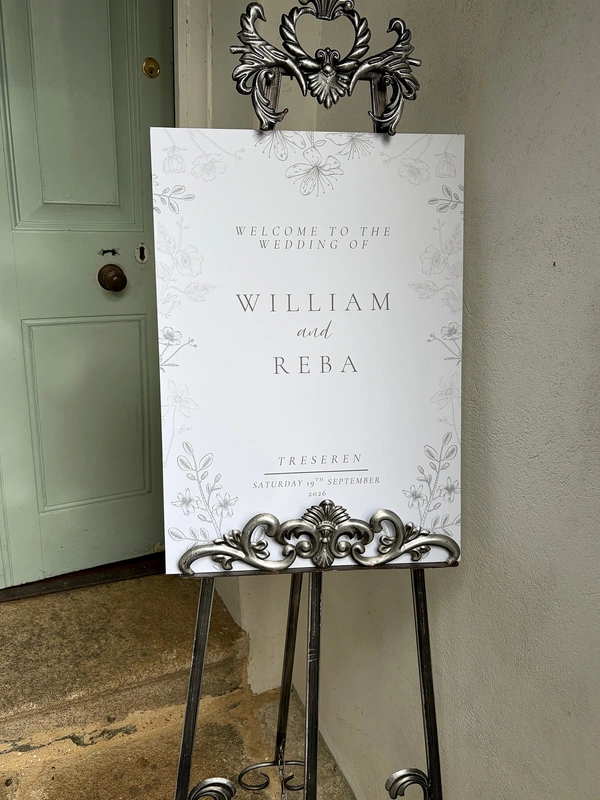 A personalised wedding welcome sign printed by Ashley Press mounted on an ornate, silver vintage style metal easel. The white sign features subtle grey floral sketches framing elegant typography that reads Welcome to the Wedding of William and Reba, along with the venue Treseren and date. It is set against an off white wall next to a pale sage green door.