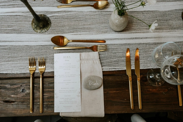 Overhead flat lay of a rustic wedding place setting set on a dark wood table with a light linen runner. In the centre, a custom wedding menu printed by Ashley Press rests on a folded linen napkin alongside a small gray pebble handwritten with Bride in white script. The setting is framed by polished gold forks, knives, and spoons, clear wine glasses, and a small ceramic bud vase with delicate white flowers.