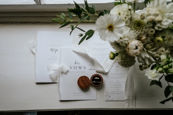 Top-down view of elegant white wedding stationery printed by Ashley Press on a rustic painted windowsill. The suite includes custom vow booklets reading A Letter to My Wife and Her Vows bound with white frayed ribbons, alongside printed invitation cards and a menu. In the centre sits an open, engraved round wooden box revealing wedding rings, next to a lush floral bouquet of white cosmos and dahlias with leafy greenery.